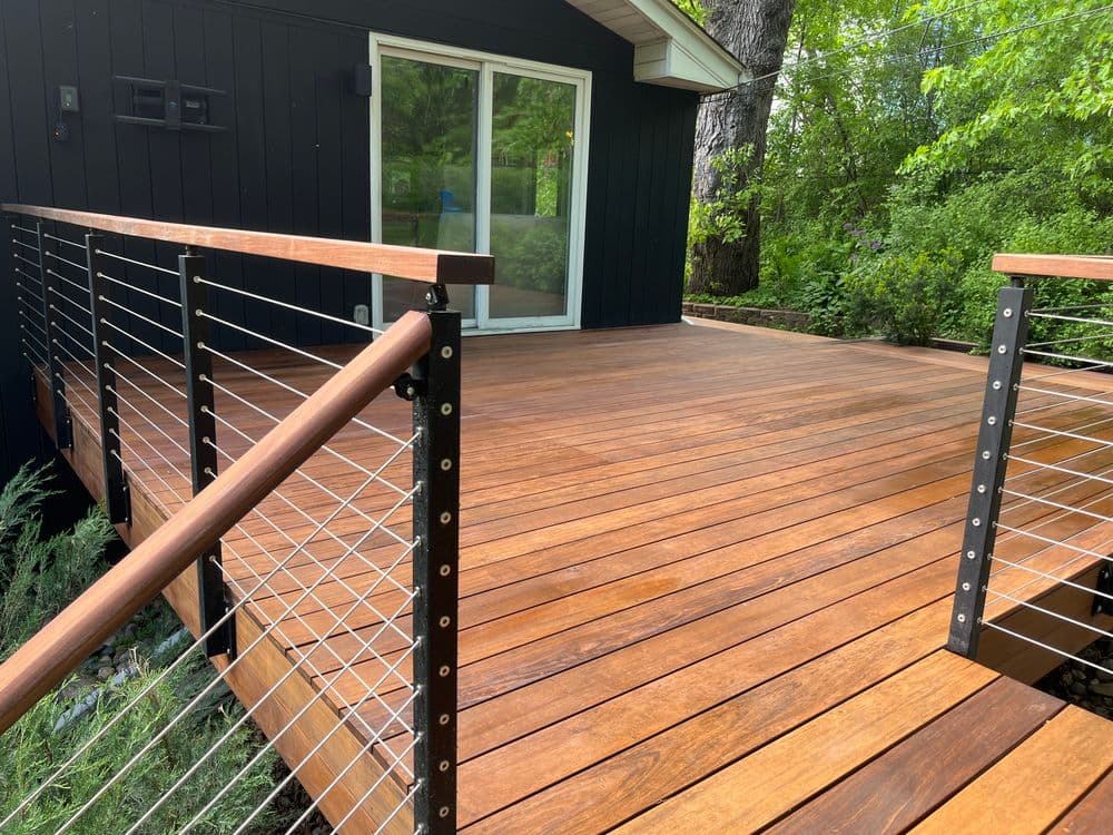 Modern wooden deck with sleek railing, surrounded by lush greenery and a contemporary house.