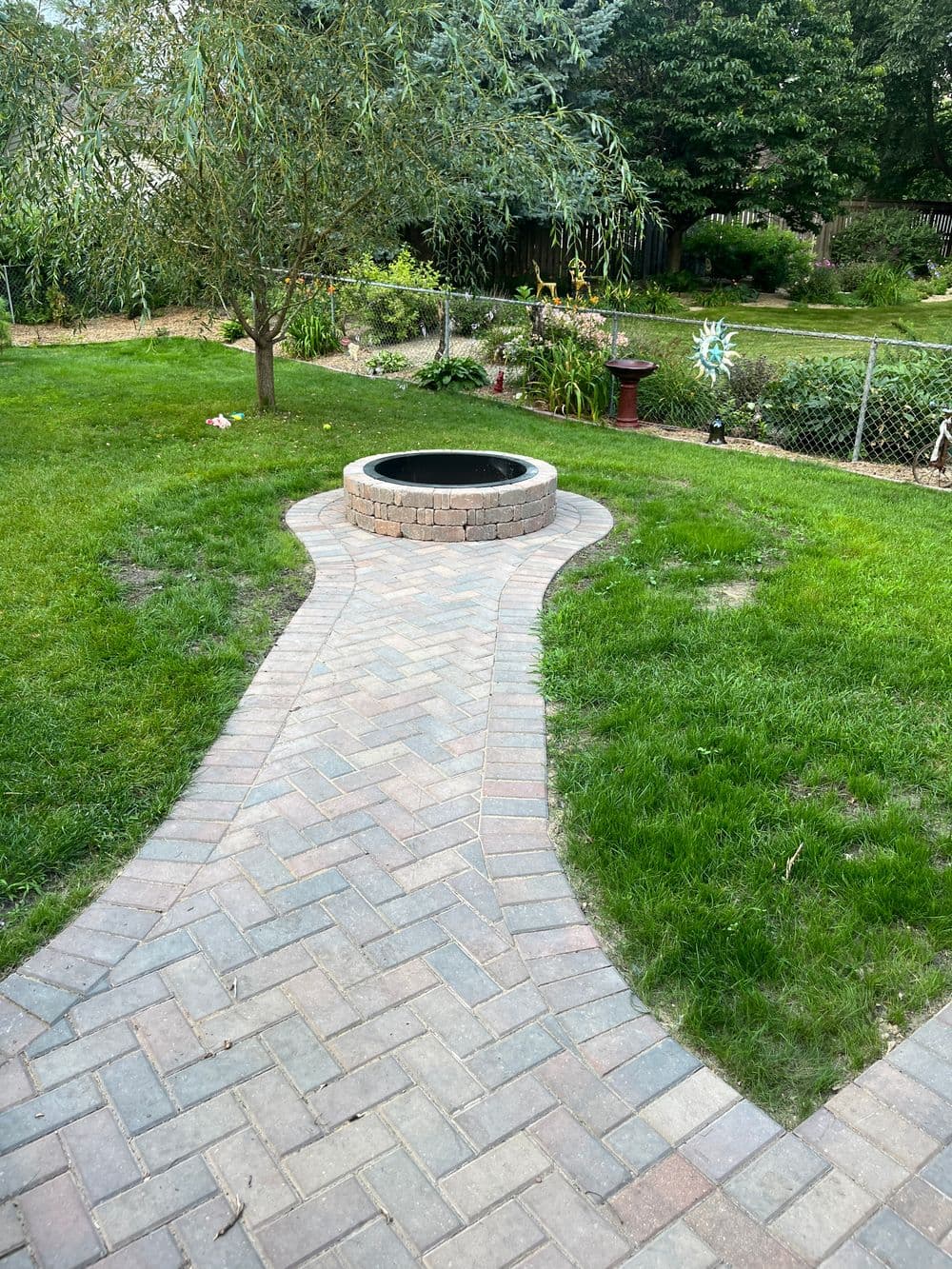 Circular stone fire pit surrounded by a paved walkway in a lush backyard garden.
