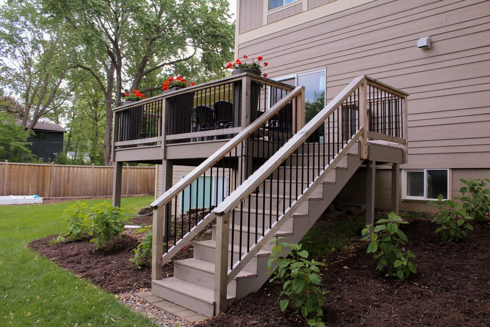 Wooden deck with railing, stairs, and flower pots beside a house in a landscaped yard.