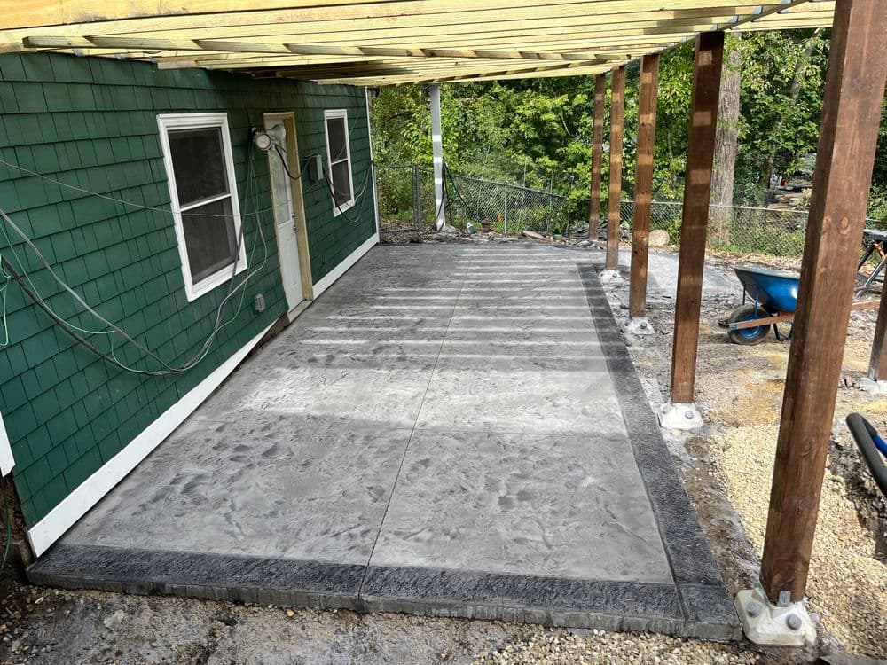 Concrete patio under wooden structure beside green shingle house, ready for outdoor use.